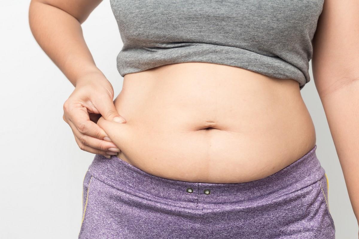 overweight woman hand pinching excessive belly fat on gray background, healthy concept
