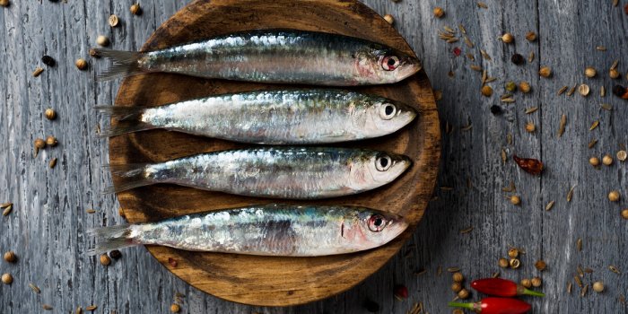 high-angle shot of some raw sardines in a wooden plate, placed on a gray rustic wooden table sprinkled with peppercorns
