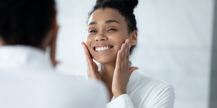 head shot happy beautiful young african american woman in white bathrobe touching face, feeling satisfied with moisturize...