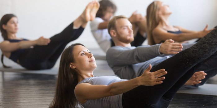 group of young sporty people practicing fitness lesson with instructor, doing warming up exercise, boat pose, working out...