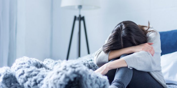 young woman in gray clothes is sitting curled up on bed at home upset brunette girl is suffering, crying depression becau...