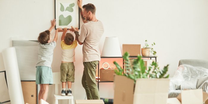 full length portrait of happy father with two sons hanging pictures on wall while moving in to new home, copy space