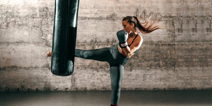 dedicated strong brunette with ponytail, in sportswear, bare foot and with boxing gloves kicking sack in gym