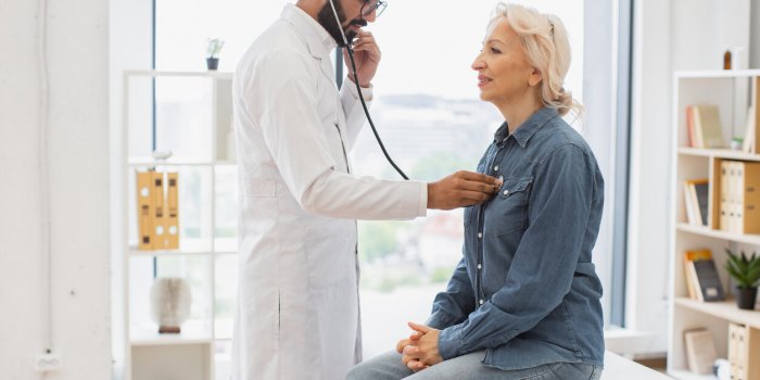 side view of focused adult man in white coat using stethoscope while elderly woman sitting on exam couch general practiti...