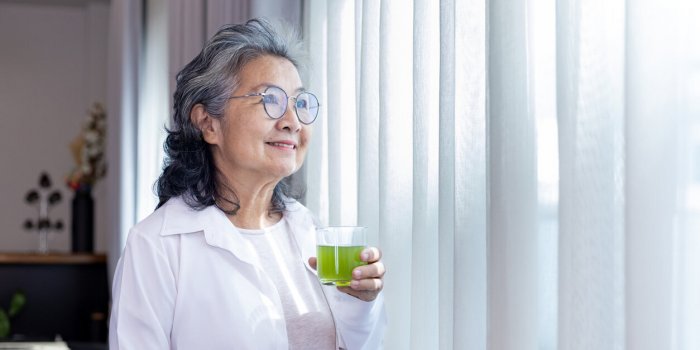 senior asian lady drinking glass of herbal supplement drink while looking out the window with copy space for nutrition bo...