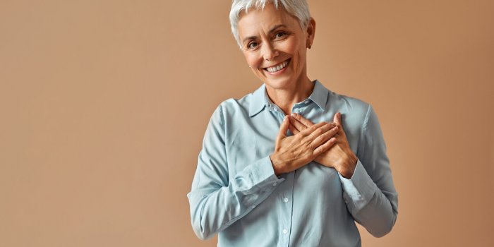 beautiful confident woman in blue blouse holding hands over heart while standing on beige background and smiling