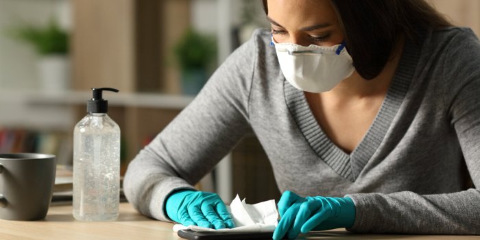 woman with gloves and protective mask disinfecting smart phone from coronavirus sitting on a desk at night at home