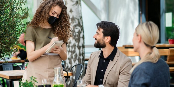 woman waitress with face mask serving happy couple outdoors on terrace restaurant