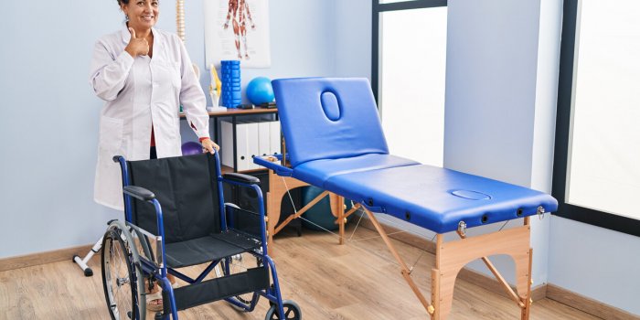 middle age hispanic woman with wheelchair at physiotherapy clinic pointing finger to one self smiling happy and proud