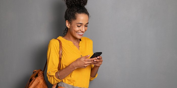 young beautiful african woman with leather backpack standing on grey wall while using smartphone stylish latin girl isola...