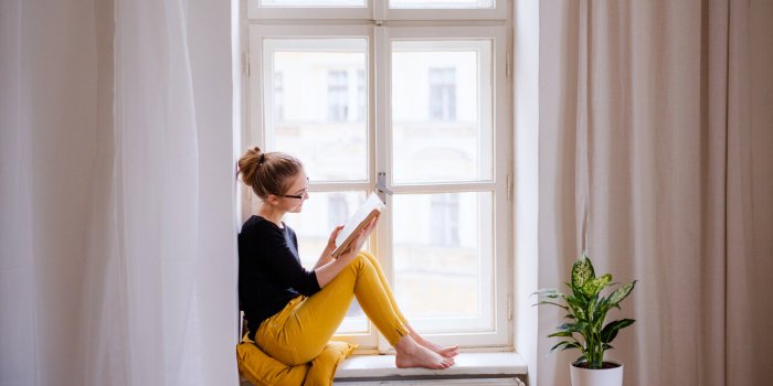 a young happy college female student with a book sitting on window sill at home, studying