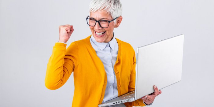 portrait of a cheerful mature woman with a laptop computer and celebrating success isolated over gray background senior l...