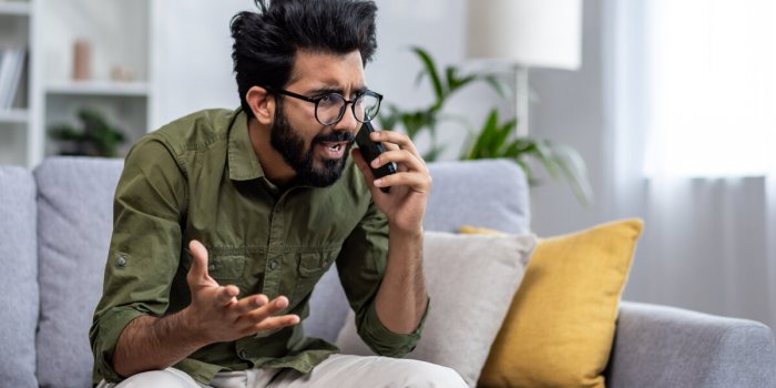 angry and nervous man talking on the phone sitting on the sofa in the living room, hispanic man yelling at the interlocut...