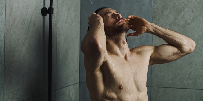 a shirtless man with a beard stands under a shower, water cascading down as he enjoys a moment of refreshment