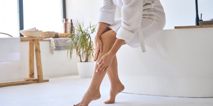 mid age adult 50s age mature woman applying varicose prevention treatment cream massaging legs sitting on bathtub wearing...