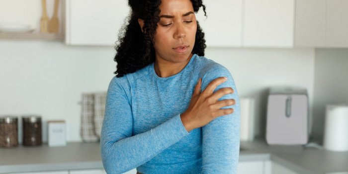 sporty black woman putting her hand on injury shoulder, having pain, standing in kitchen interior at home, resting after ...