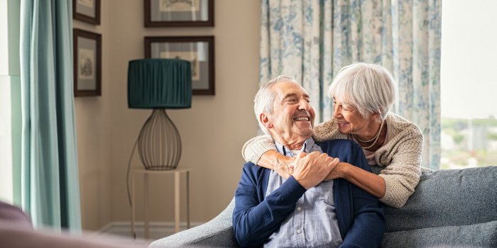 happy senior woman embracing her husband at home while laughing together smiling wife hugging old man sitting on couch fr...