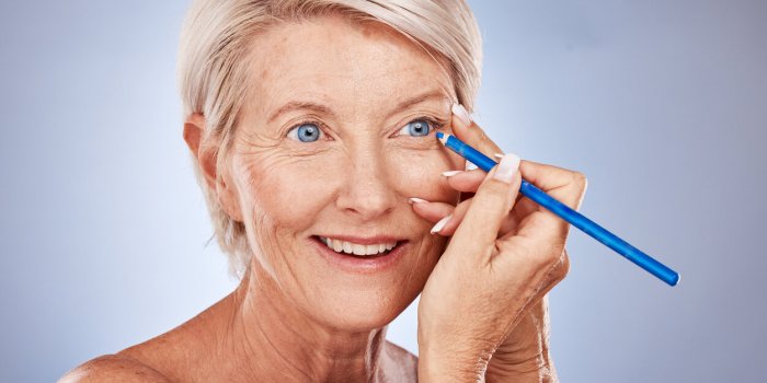 face, beauty and eyeliner with a senior woman in studio on a gray background to apply cosmetics makeup, beautiful and eye...