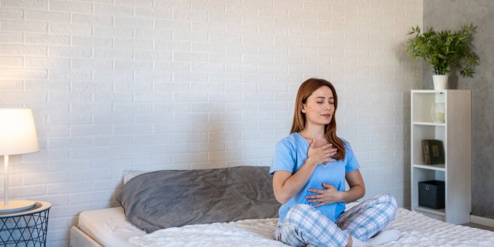 young woman with her eyes closed practices diaphragmatic breathing exercises on her bed in her bedroom, promoting relaxat...